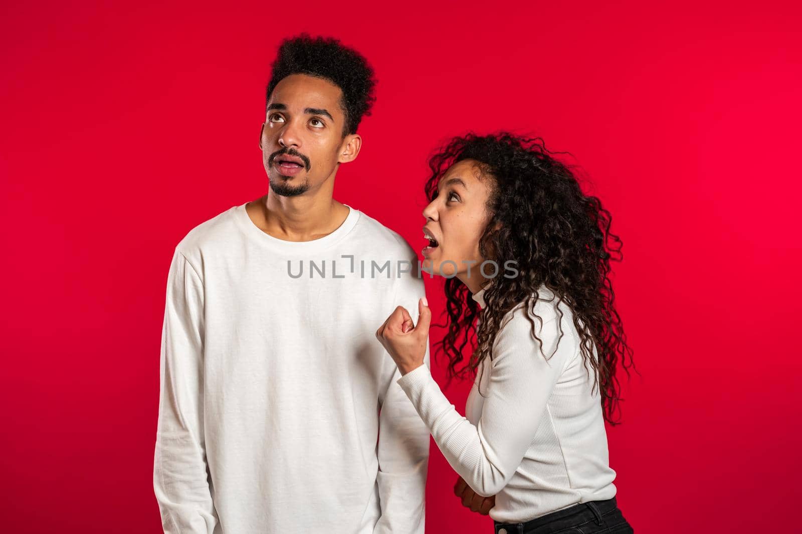 Young african woman emotionally screaming at her husband or boyfriend on red background in studio. Bored man rolling his eyes. Concept of conflict, problems in relationships