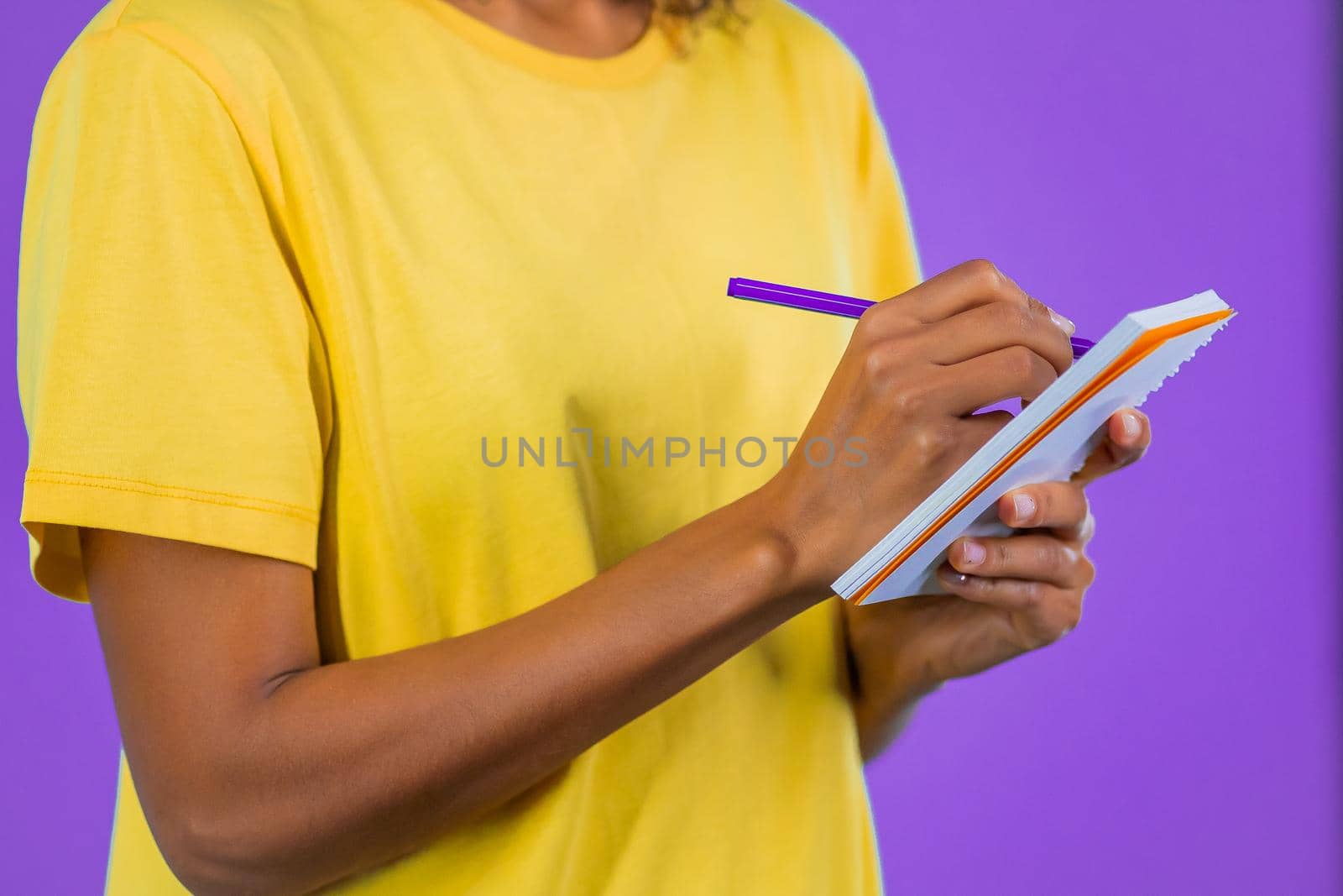 YHands of young woman making notes in planner, african lady holding pen. She writes future plans and to-do list in notebook for week or month. Keeping personal diary on purple studio background. by kristina_kokhanova