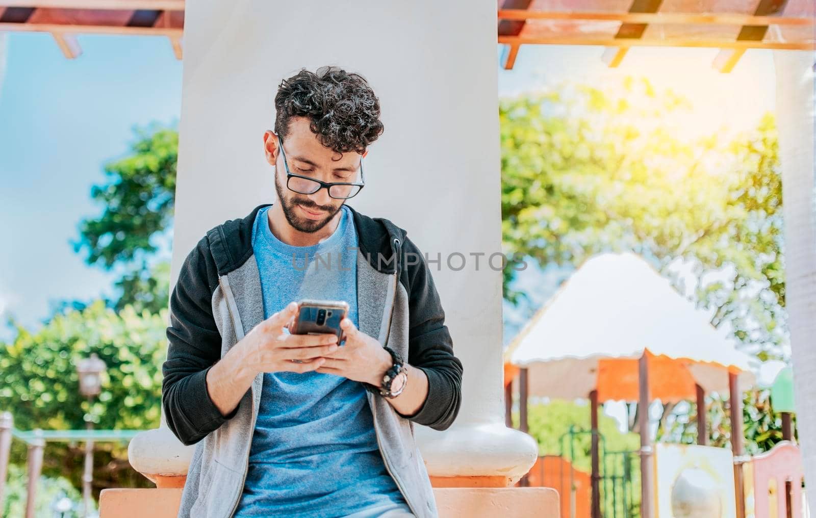 Handsome guy in glasses leaning on a wall chatting on his cell phone, Teenage male leaning on a wall sending a text message, Smiling handsome man using a phone leaning on a wall in a park by isaiphoto