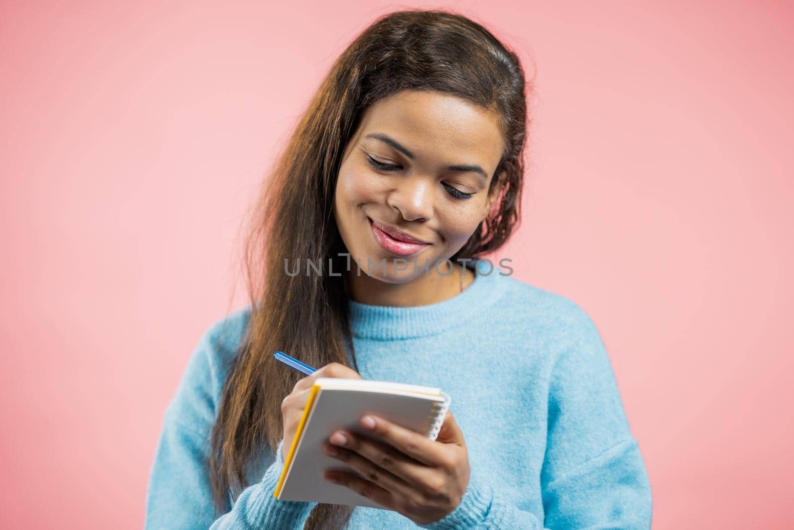 African woman making notes in planner, mature lady holding pen. She writes future plans and to-do list in notebook for week or month. Keeping personal diary on pink studio background