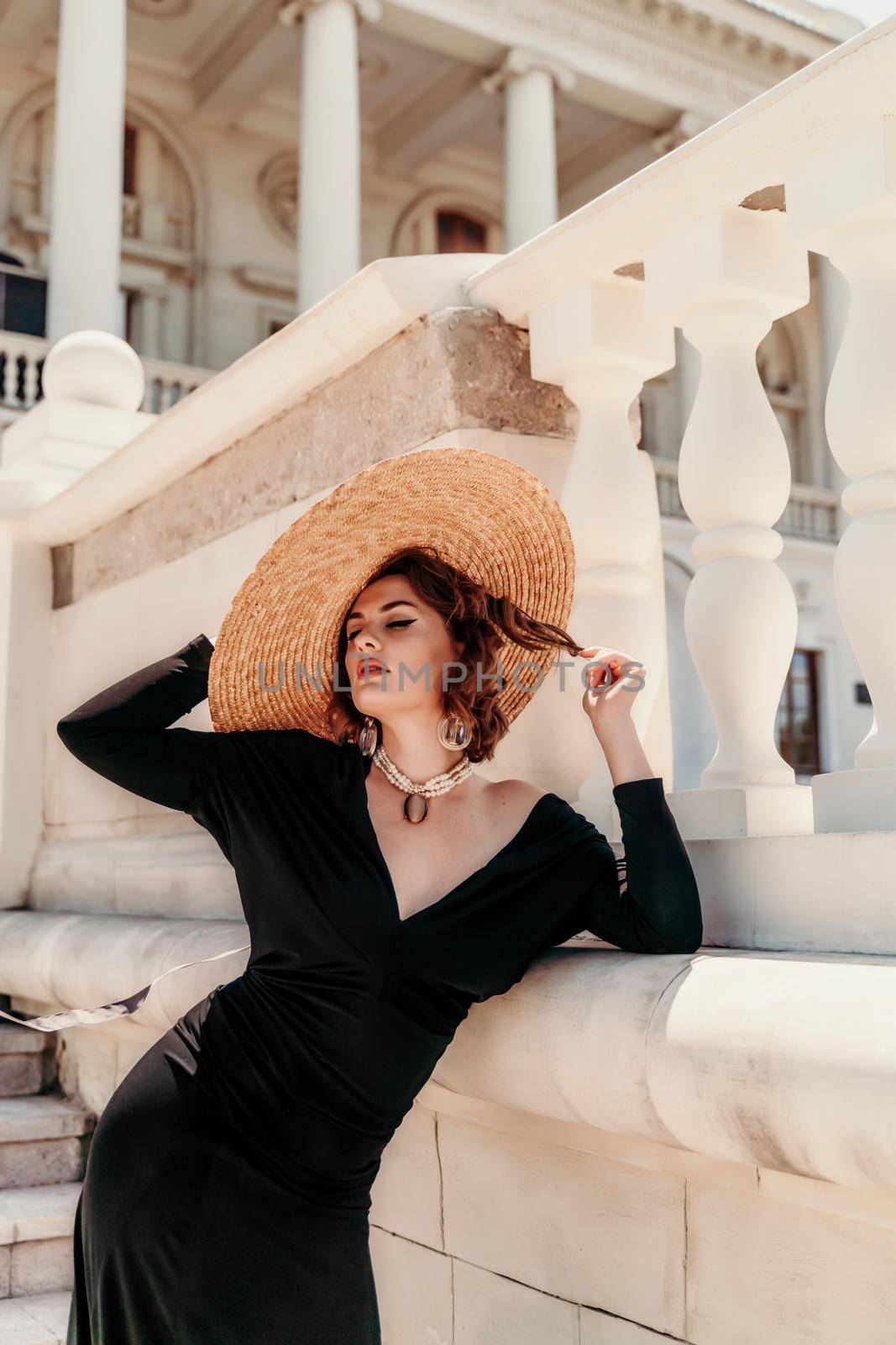 Fashion photo of a beautiful model in an elegant hat and black dress posing against the backdrop of a building with columns