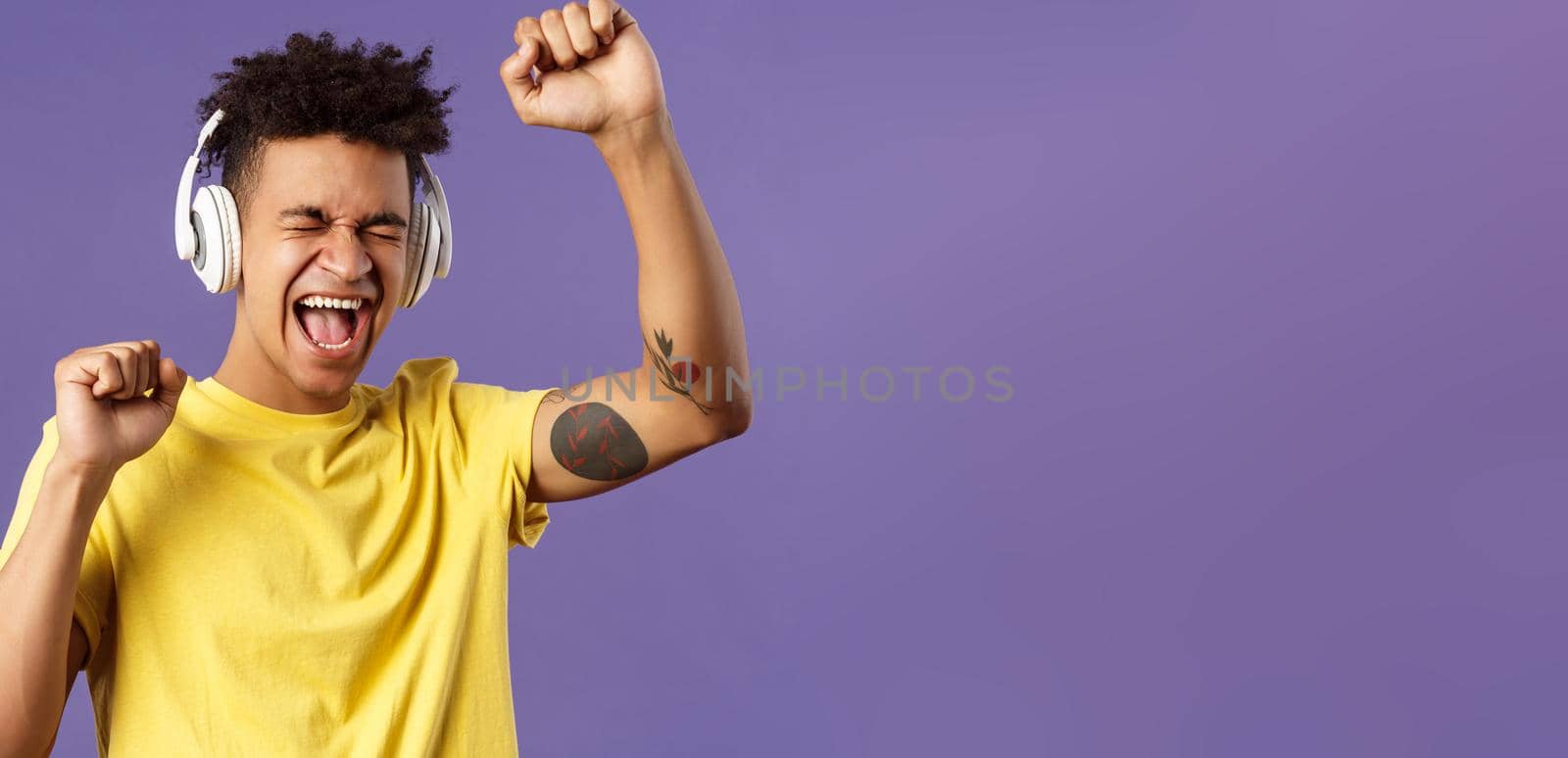 Close-up portrait of cheerful, happy young dancing guy lift hand up singing along, close eyes and smiling upbeat as listening awesome song in headphones, enjoying music, purple background by Benzoix