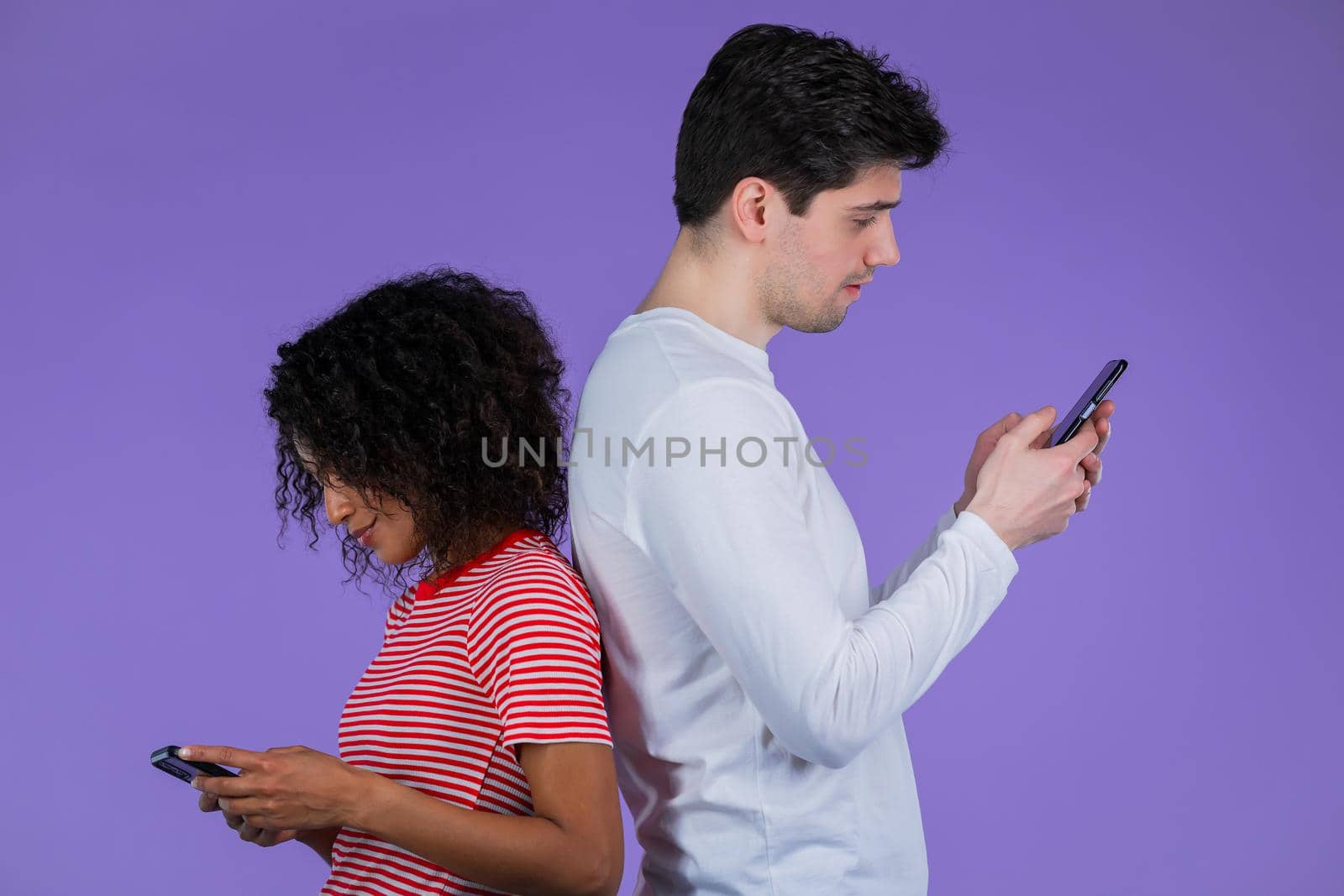 Interracial couple stand with their backs to each other and print messages or surf Internet from mobile phones. Technology, distancing, smartphone, social networks concept.