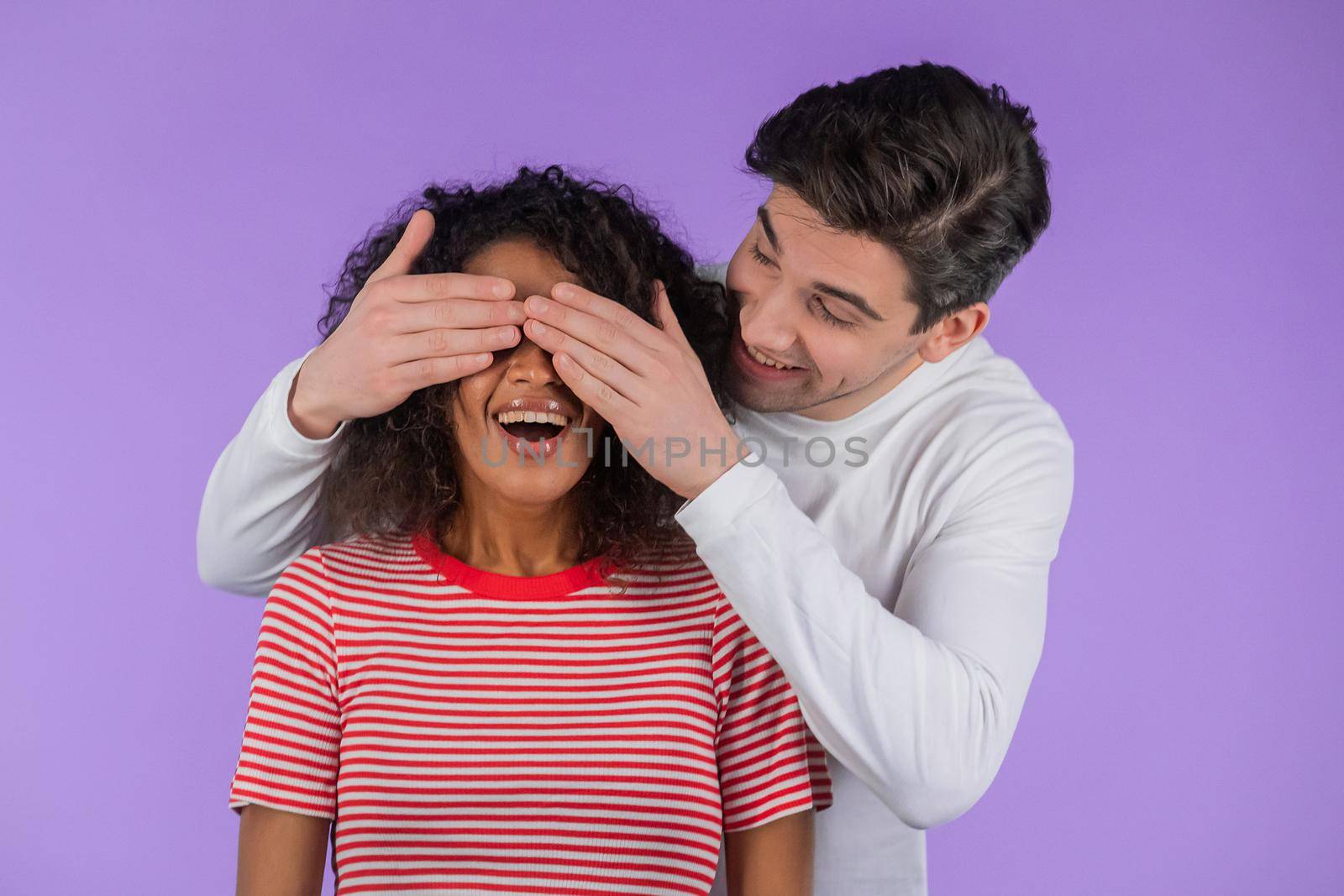 White man closes eyes of his beloved african american girlfriend before surprise her. interracial couple on purple studio background. Love, holiday, happiness concept. High quality photo