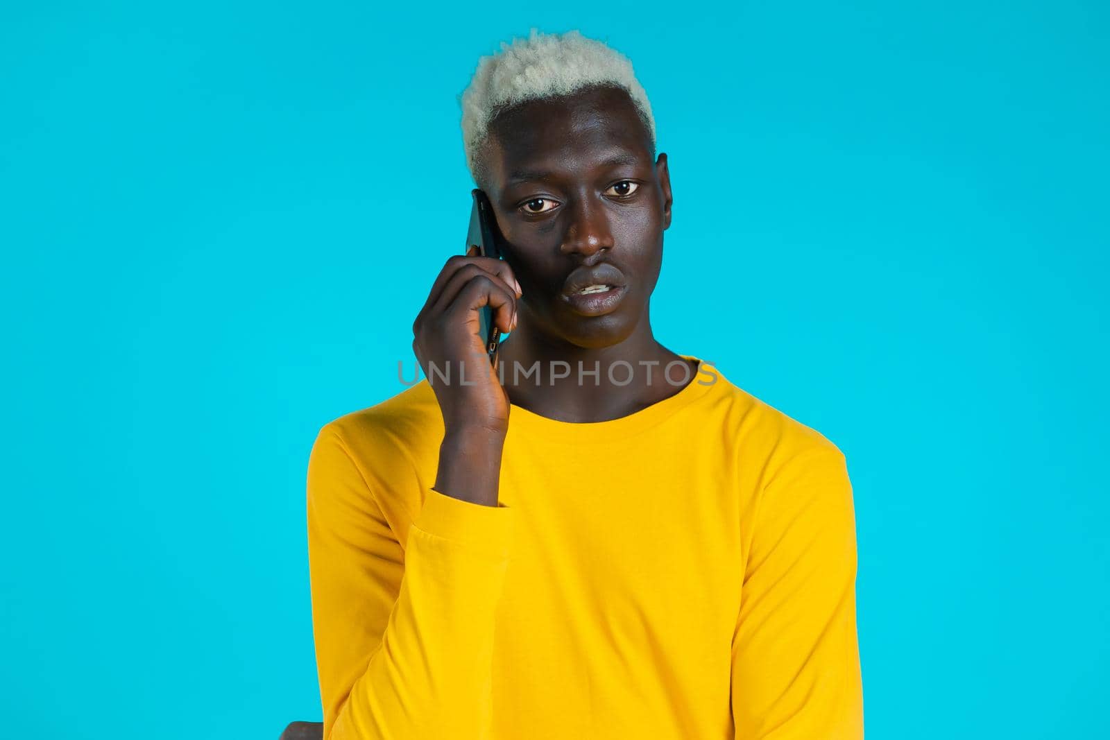 Young afro man speaks serious on phone. Guy holding and using smart phone. Blue studio background. by kristina_kokhanova