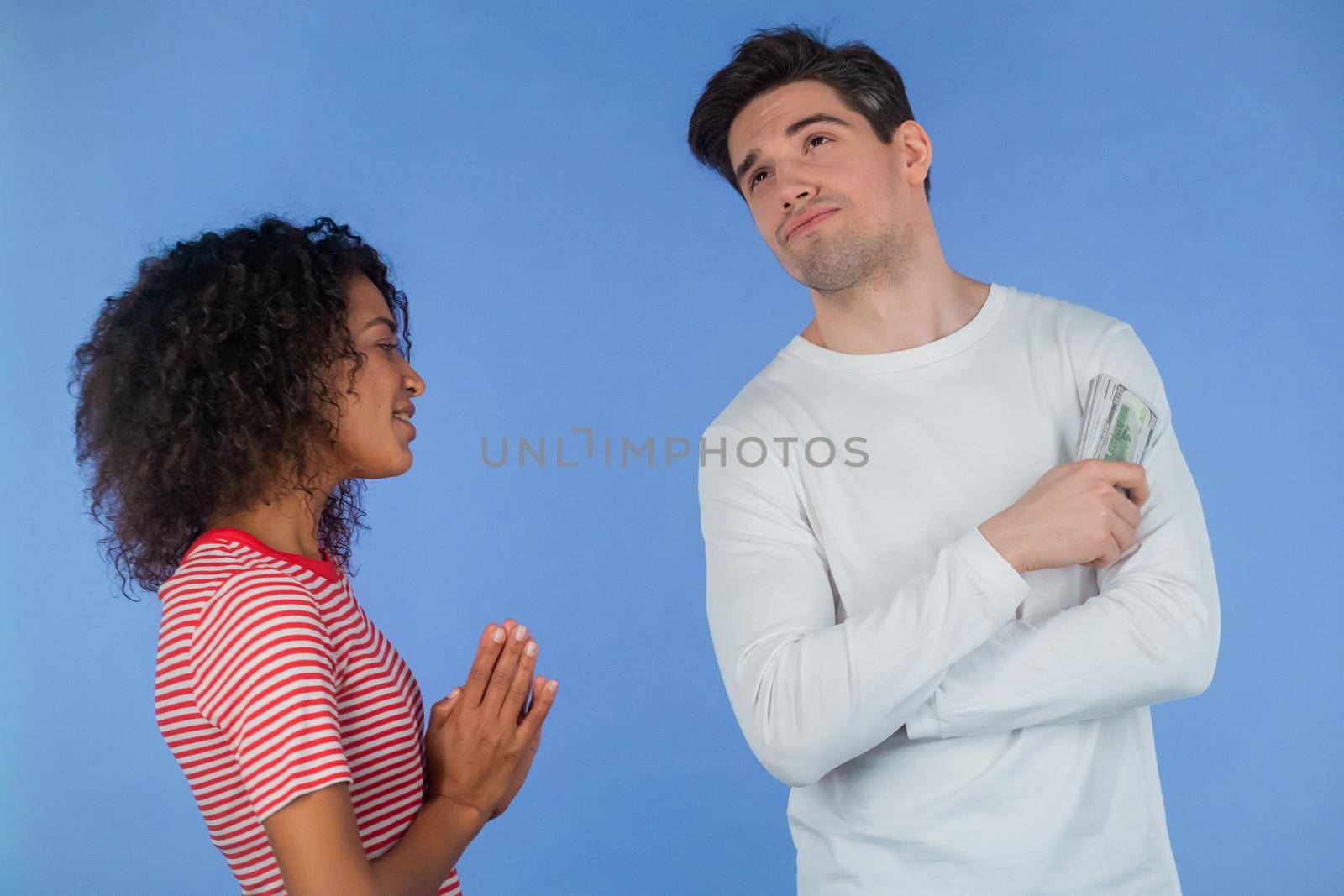 African girl asks her boyfriend for money for her whims, beauty salon and shopping. Husband reluctantly counts her dollar bills. Interracial couple concept. Blue studio background. High quality photo