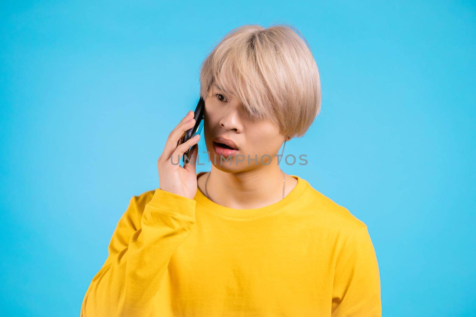Young surprised asian man speaks with smile on phone. Guy with dyed blond hair holding and using smartphone, questioning facial expression. Blue studio background