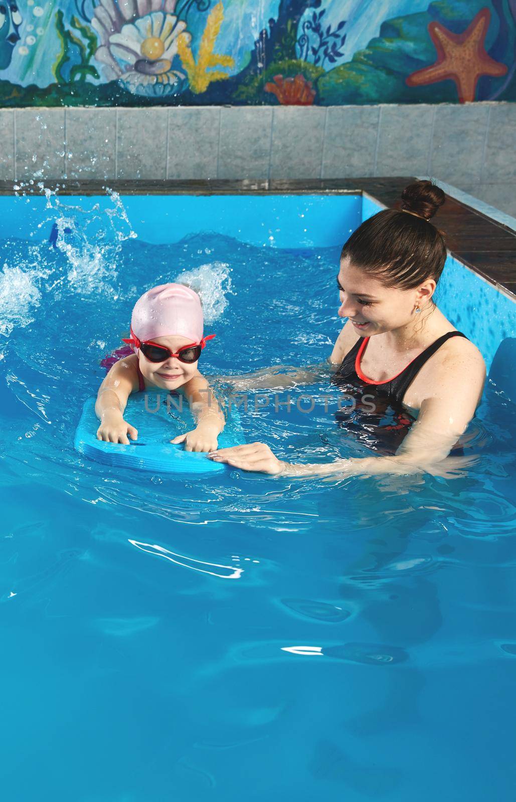 Little girl learning to swim in indoor pool with pool board and trainer by Mariakray