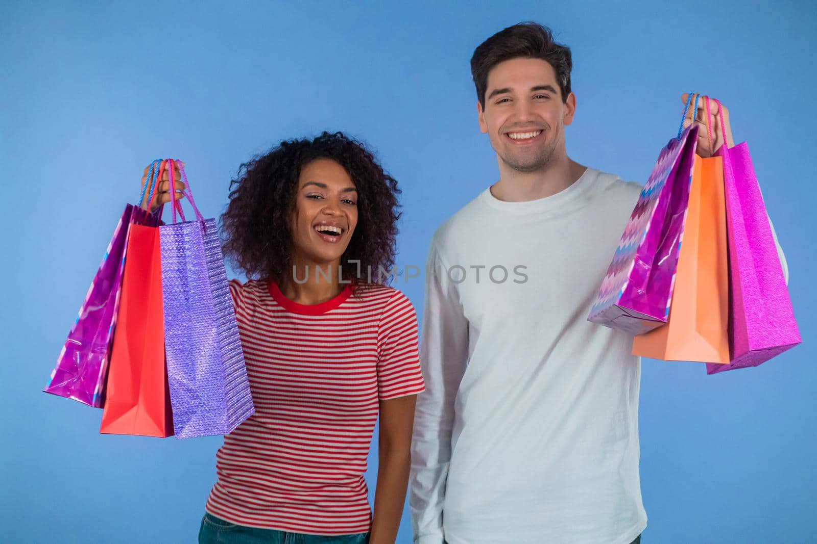Interracial shopaholic couple holds shopping paper bags on blue studio background. White man and african woman bought presents on sales with discounts in center after quarantine. High quality photo