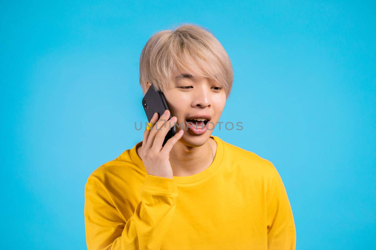 Young asian man speaks with smile on phone. Guy with dyed blond hair holding and using smartphone. Blue studio background. by kristina_kokhanova