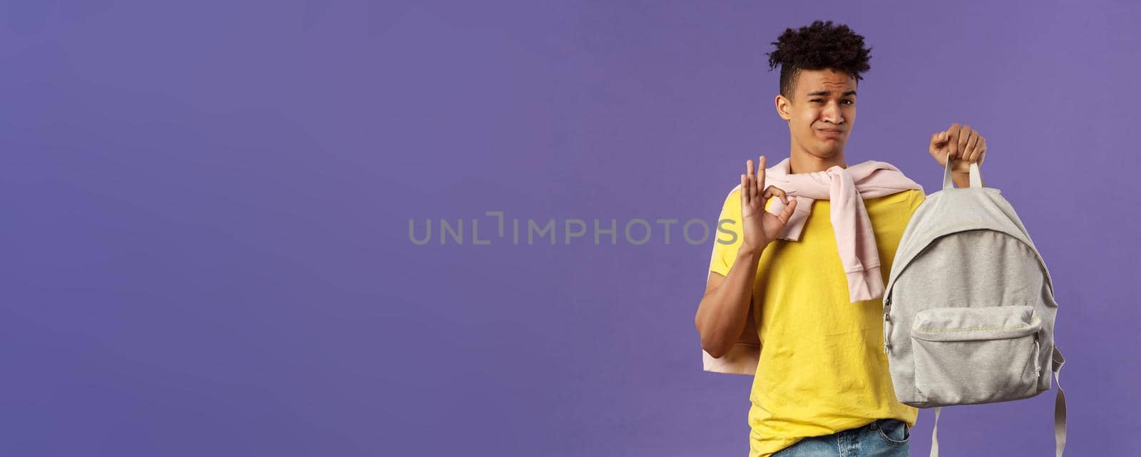 Good but not the best. Portrait of disappointed, unsatisfied young teenage student, guy grimacing without enthusiasm show okay, received new ugly backpack, standing purple background.