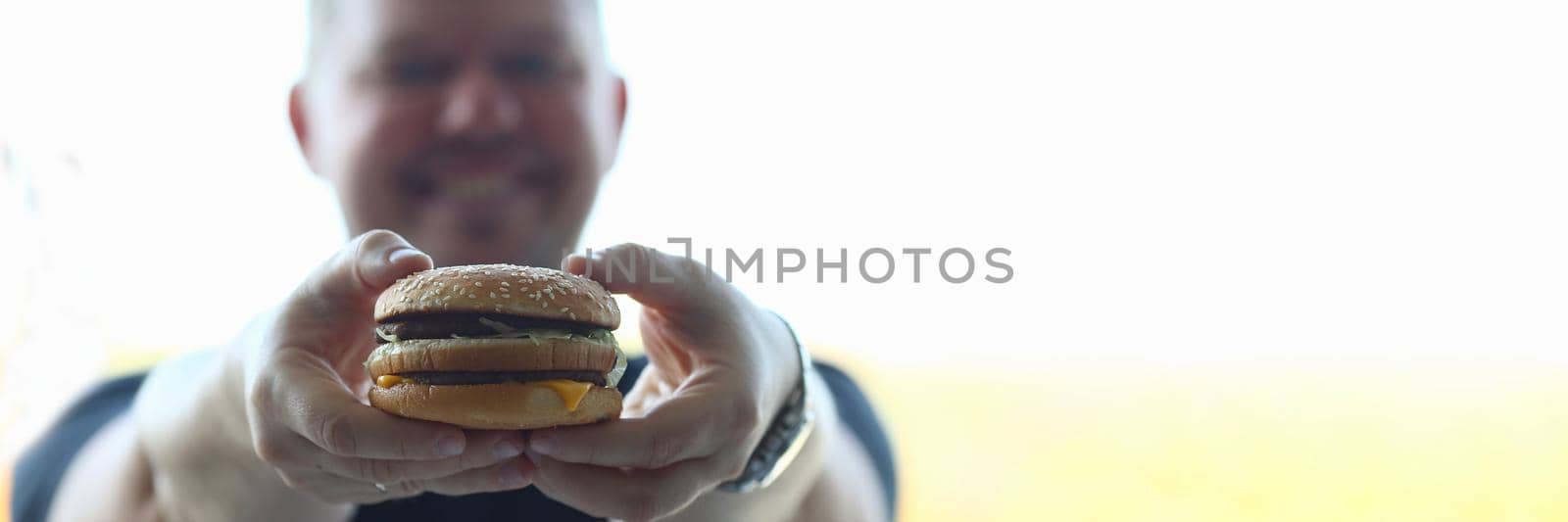 Young man holds hearty hamburger in hands. Fast food benefit and harm concept
