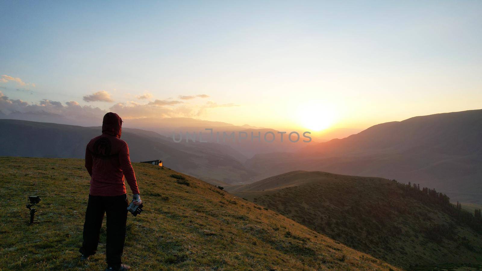 A guy in the mountains is watching the sunset. An epic orange-red sunset is reflected on small clouds and green hills. Bushes and forest grow in places on mountains. The guy is standing with his back