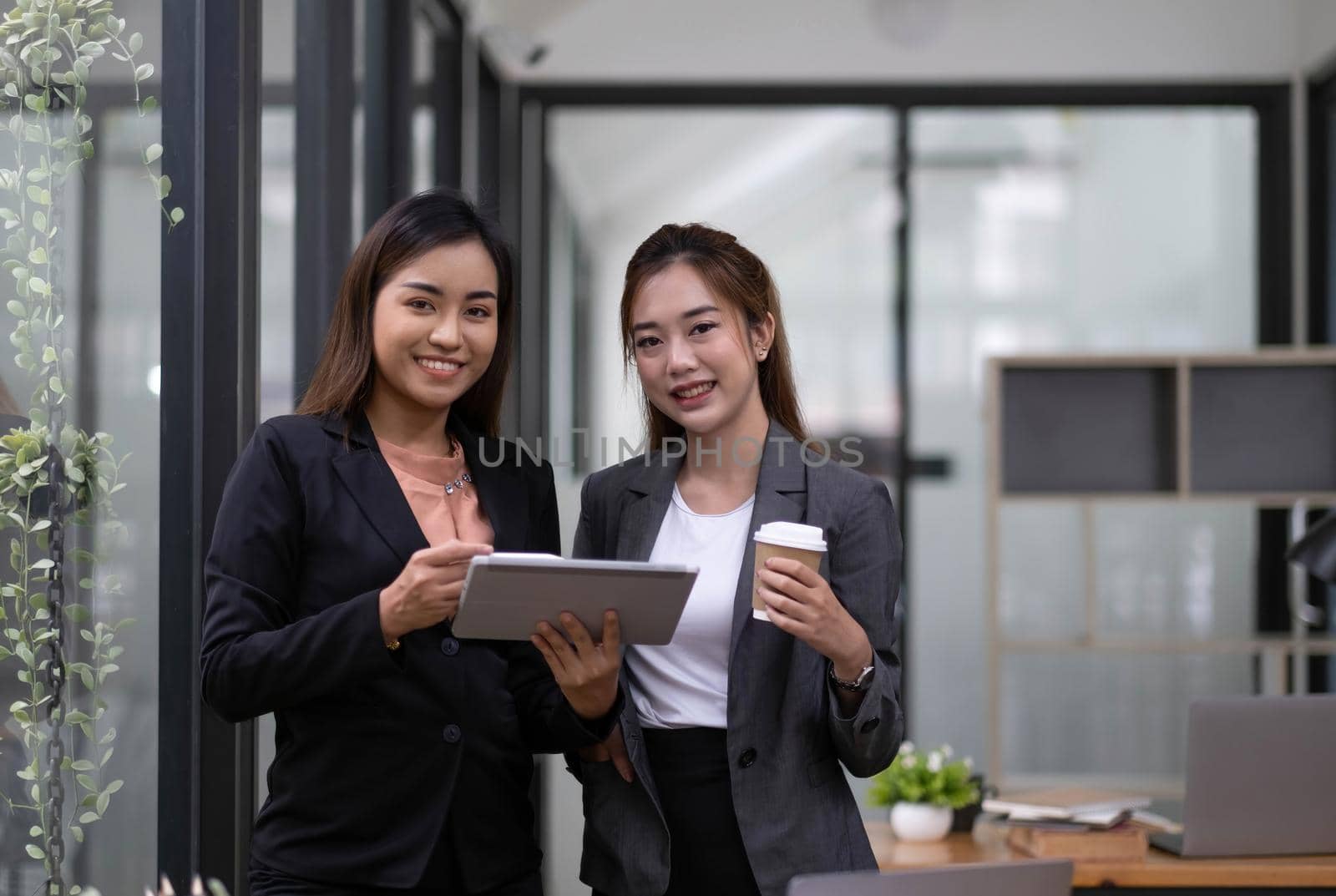 Two beautiful Asian businesswomen standing using digital tablet consulting and analyzing information in office work. by wichayada