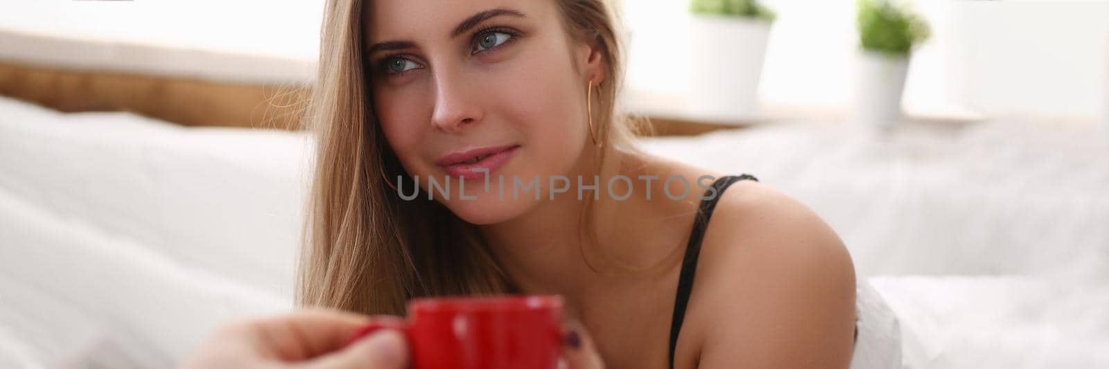 Smiling blonde is drinking cup tea with coffee early in while lying in bed by kuprevich