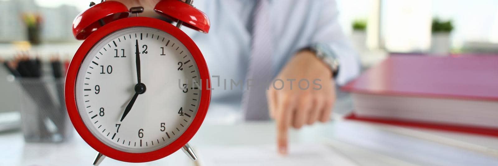 Man hand on red alarm clock stands at desk in office showing seven o'clock AM PM by kuprevich