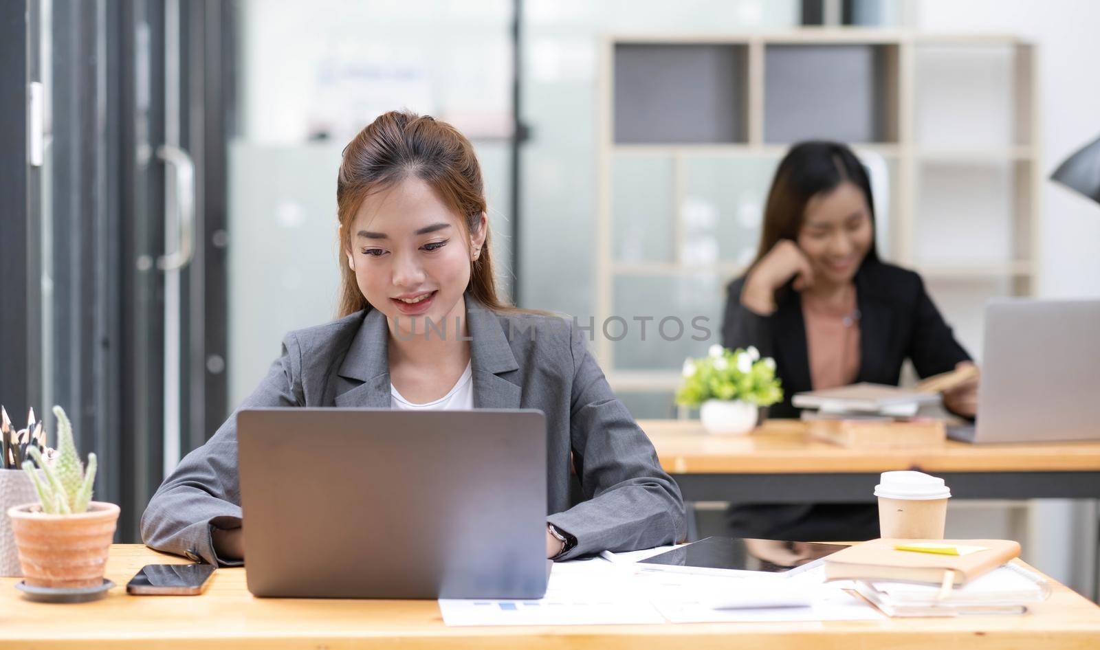 Two business women are working on a notebook computer, holding a pen and looking at the screen in office. by wichayada