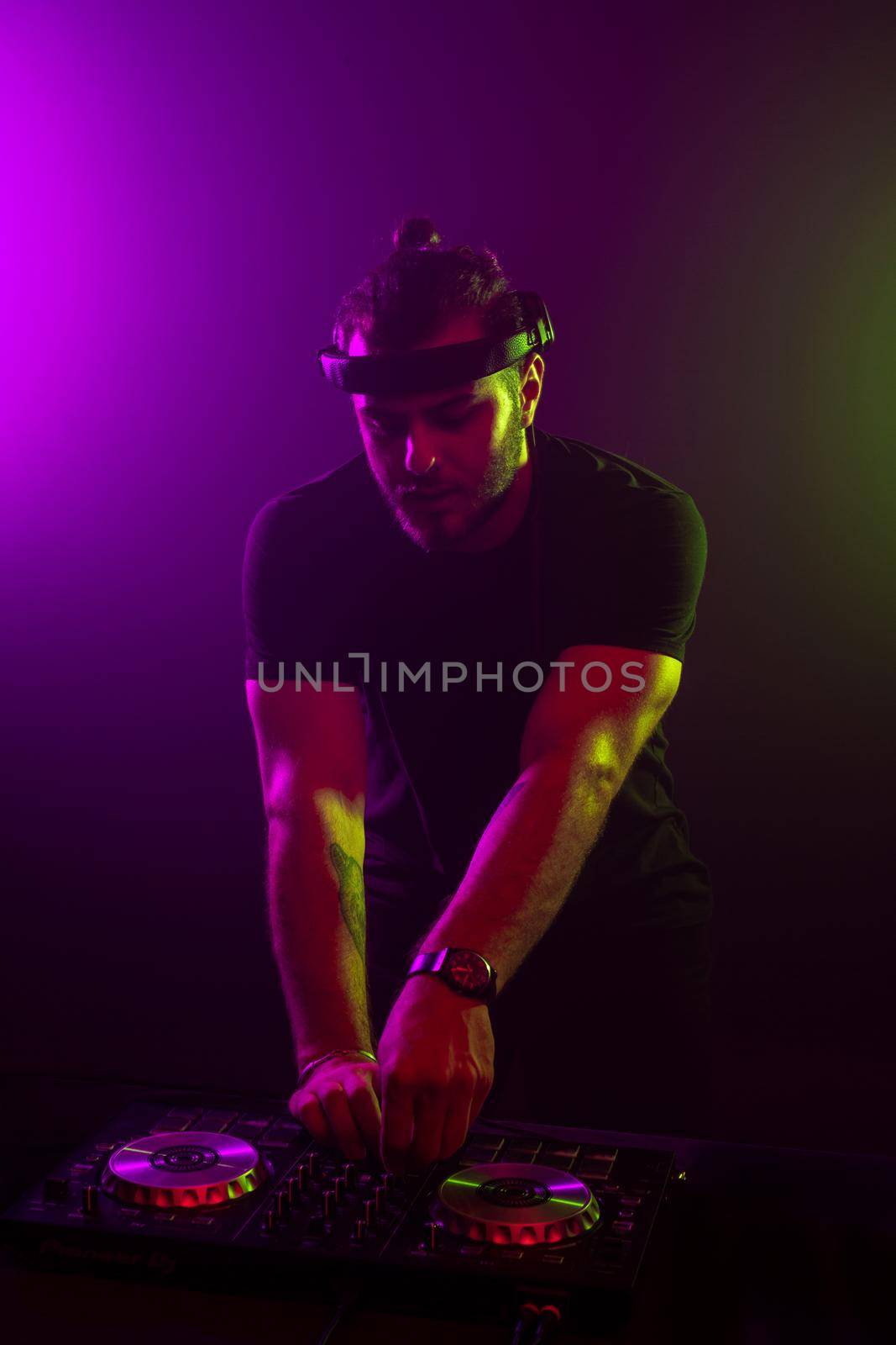 DJ playing music at mixer on colorful foggy background. A fashionable, handsome young man in a black T-shirt is behind the DJ's console. Studio shot