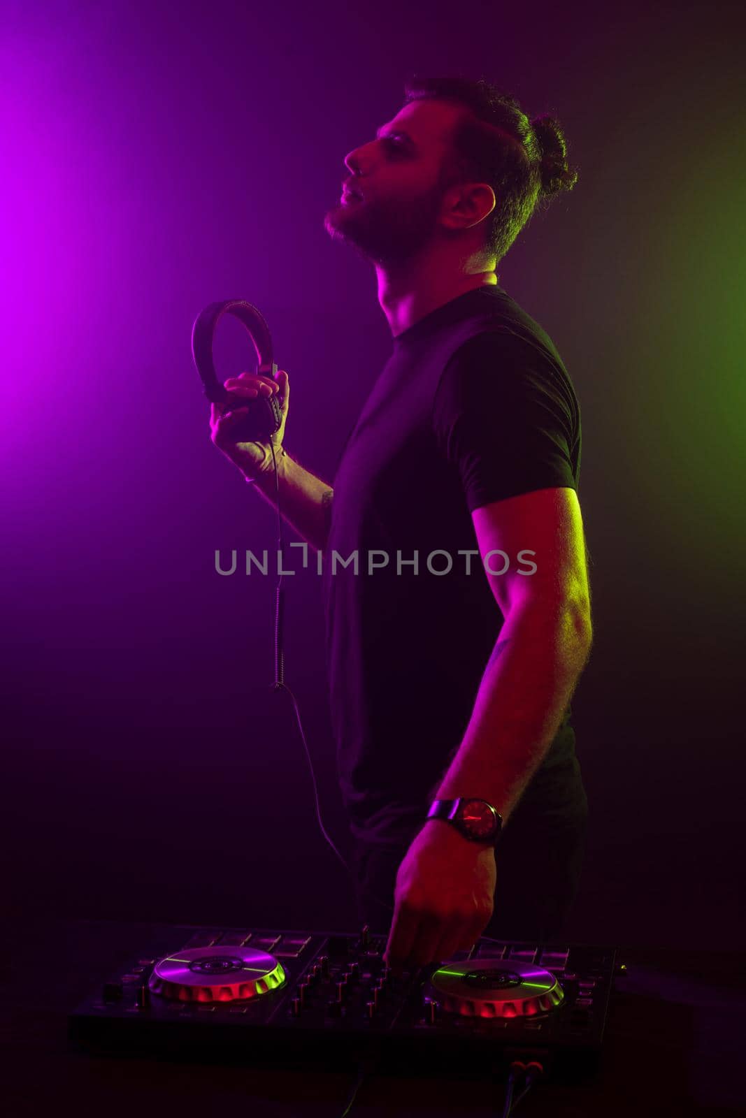 DJ playing music at mixer on colorful foggy background. A fashionable, handsome young man in a black T-shirt is behind the DJ's console. Studio shot