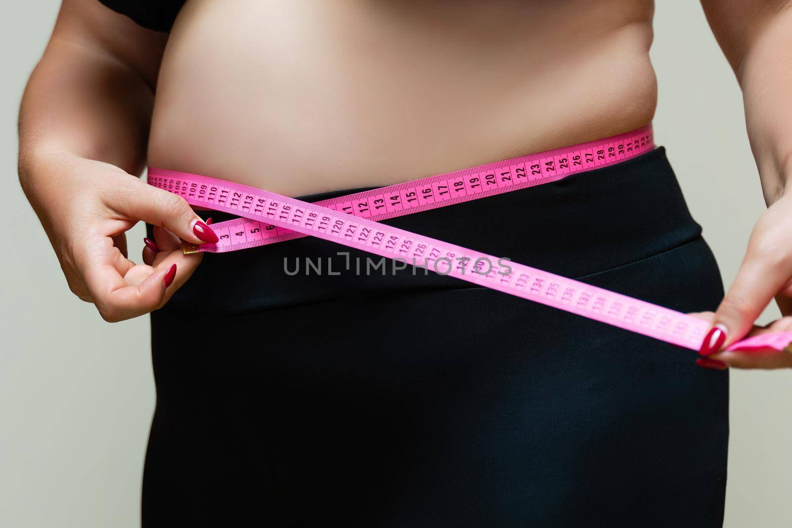 Cropped view of overweight woman measuring waist with tape measure at home, close up. Unrecognizable European woman checks result of weight loss diet or liposuction indoors by Matiunina