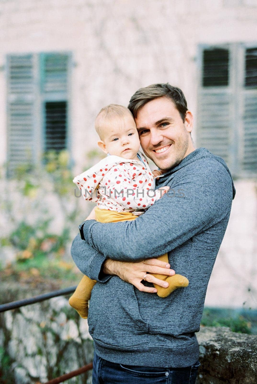 Smiling dad stands at the wall of the house hugging the baby in his arms. High quality photo