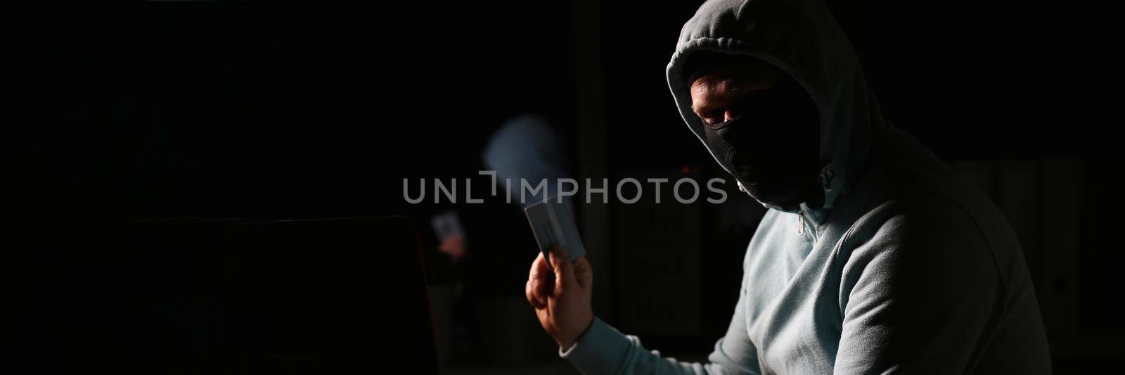 Hacker in dark jacket looking at laptop screen in dark room is holding bank card by kuprevich