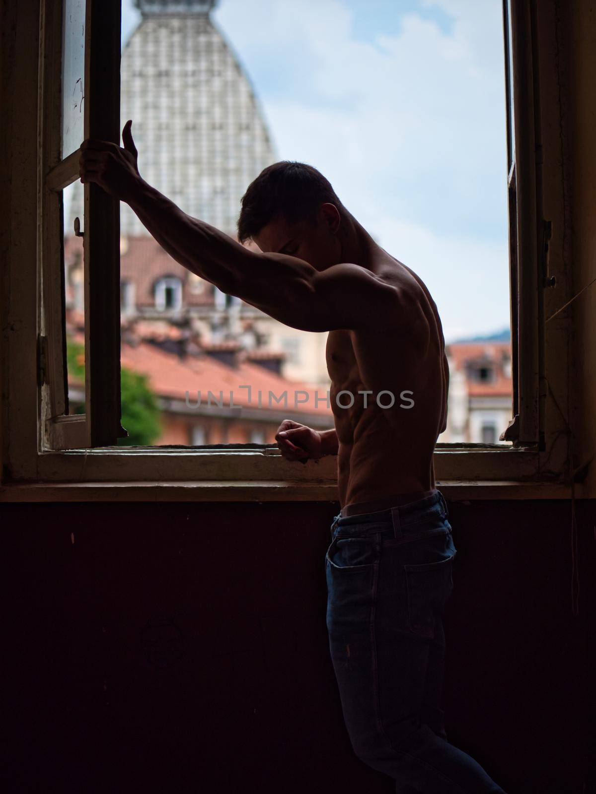 Portrait of sexy shirtless muscular man next to window curtains during the day, wearing only jeans, looking out