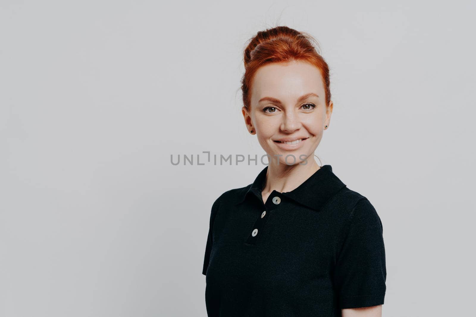 Gorgeous confident young 30s woman with red hair in bun posing isolated over grey studio background, dressed casually in black t-shirt, looking cheerfully at camera and expressing confidence