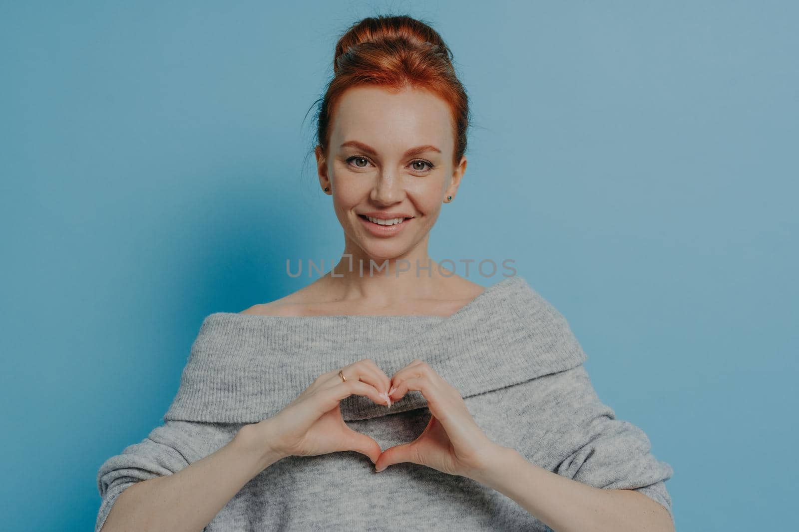 Kind smiling red haired woman showing heart gesture on chest isolated on blue wall background by vkstock