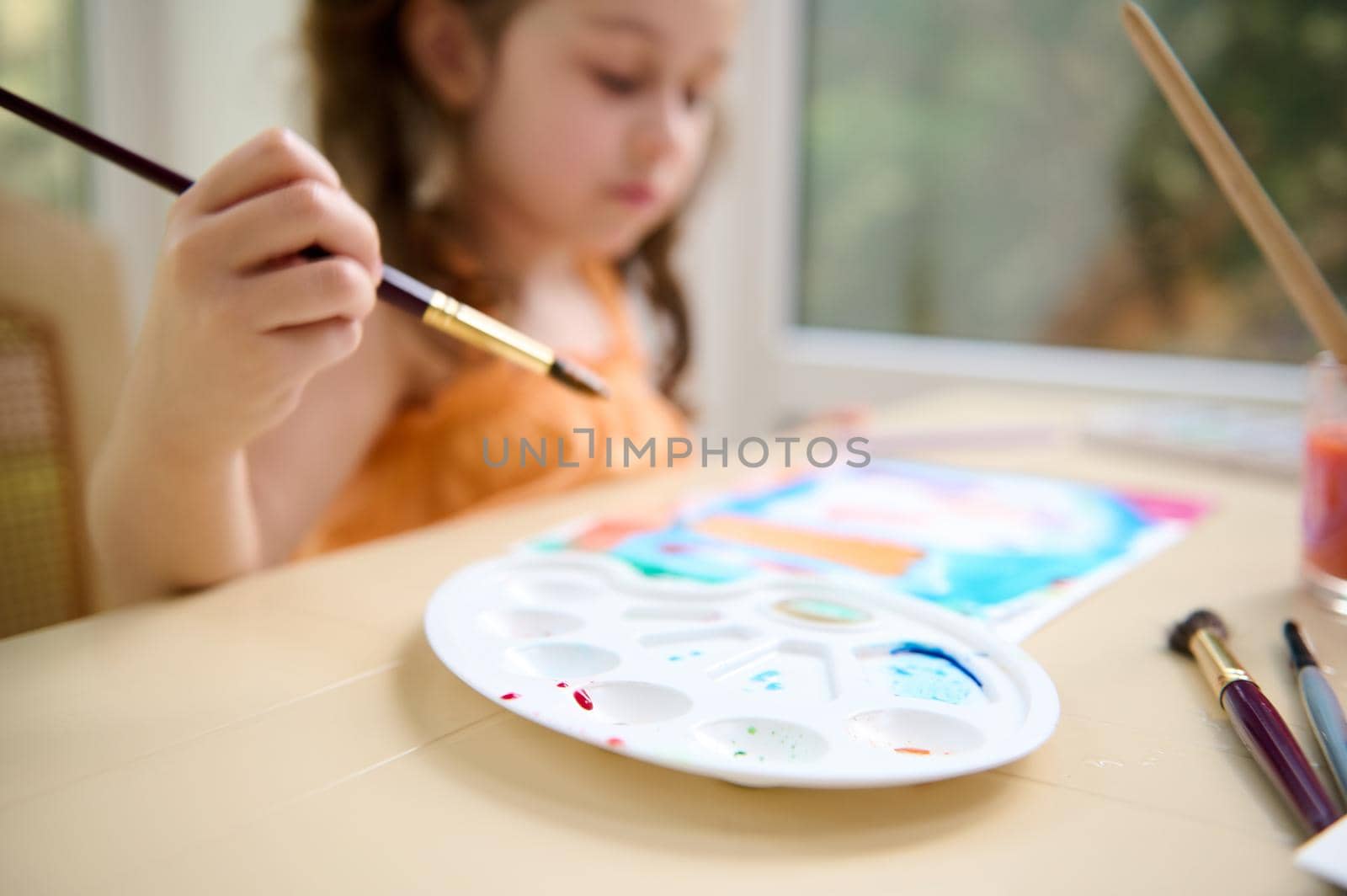 Focus on white plastic palette for mixing paints on the table on the blurred background of a little girl holding a paintbrush and painting with watercolour paints. Art master class. Child hobby