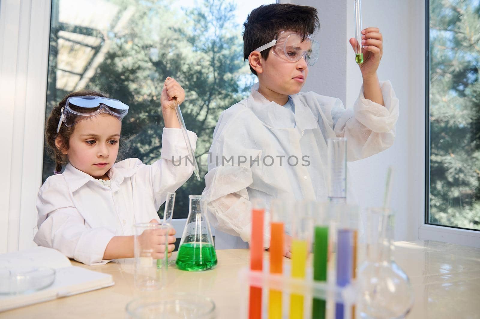 Diverse kids, teenage boy and preschooler girl in white laboratory coats and protective eyeglasses, doing biochemistry research in chemistry class. Test tubes, flasks with chemicals on the foreground