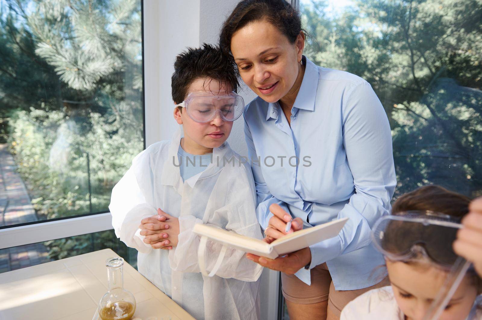 Smart kid in white lab coat and safety goggles doing experiments while standing next to chemistry teacher by artgf