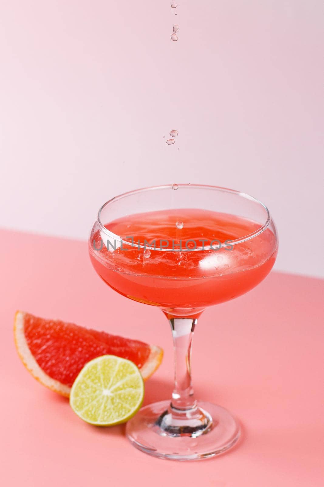 Fresh grapefruit juice with drops falling into a glass on a pink background with grapefruit slices and lime on a pink background. Copy space