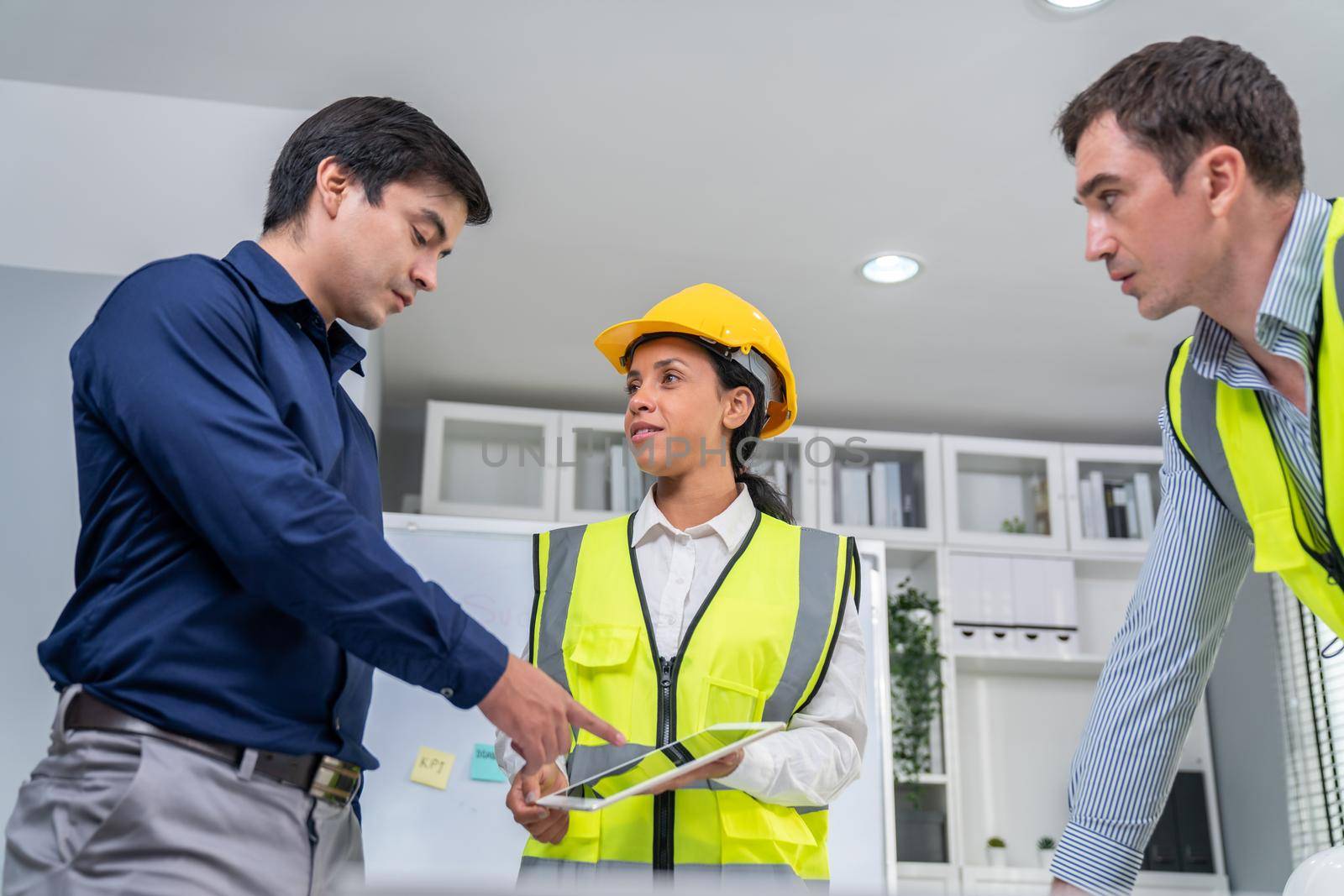 A team of competent engineers wearing safety equipment is working on blueprints with a tablet while also discussing with the investor in the office.