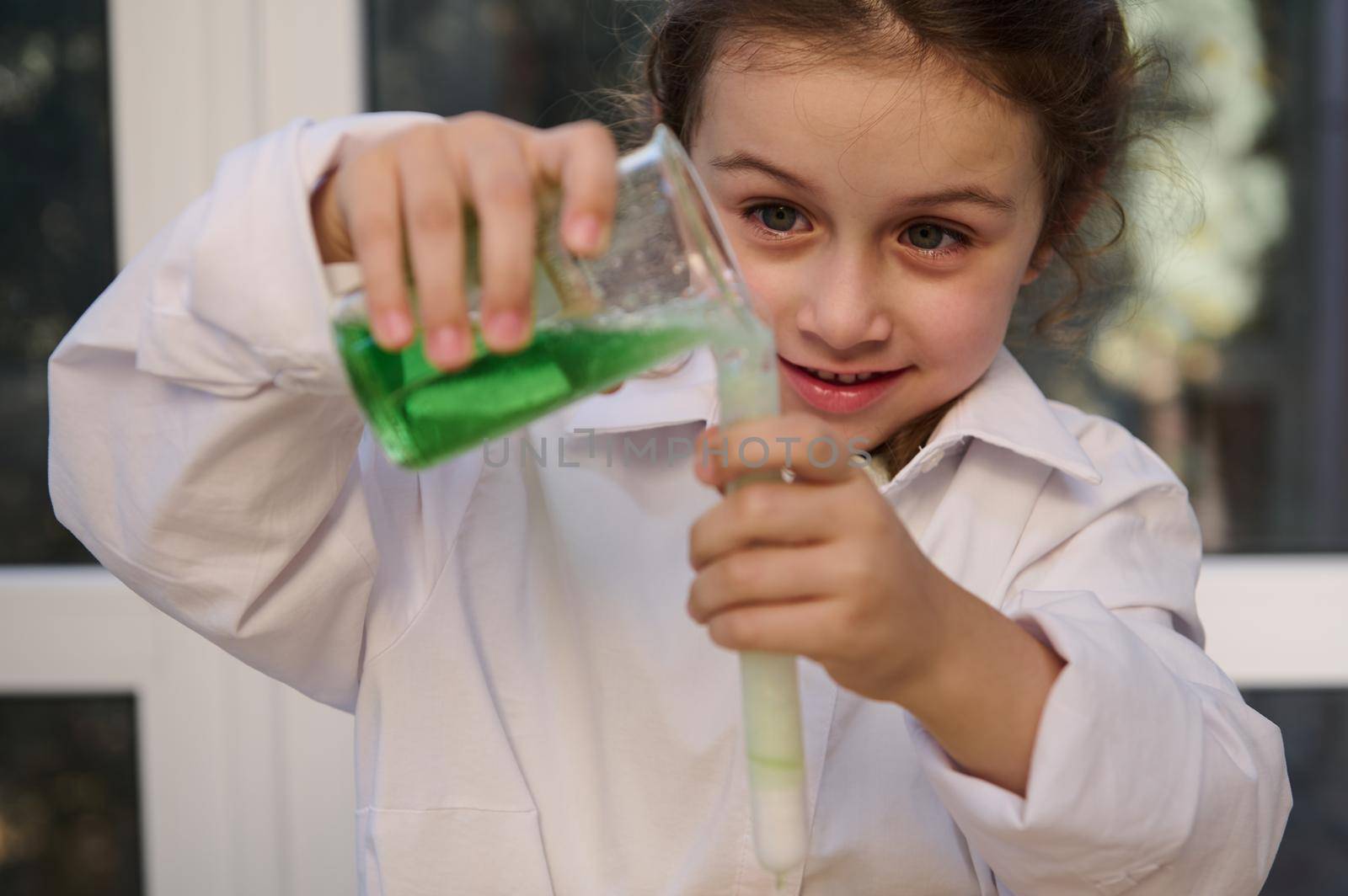 Homeschooling Caucasian little girl in white lab coat learning about science, pouring reagent solution from flask into a test tube, watching fascination going on chemical reaction. Learning chemistry
