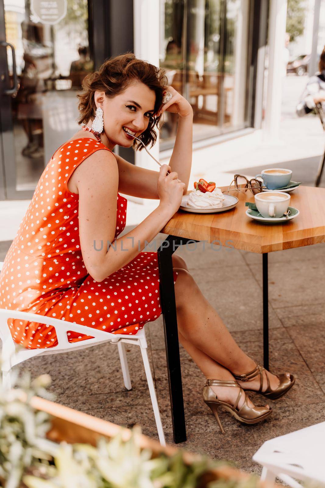 Charming woman in a restaurant, cafe on the street. She sits at the table and eats a cake with a fork. Dressed in a red sundress with white polka dots