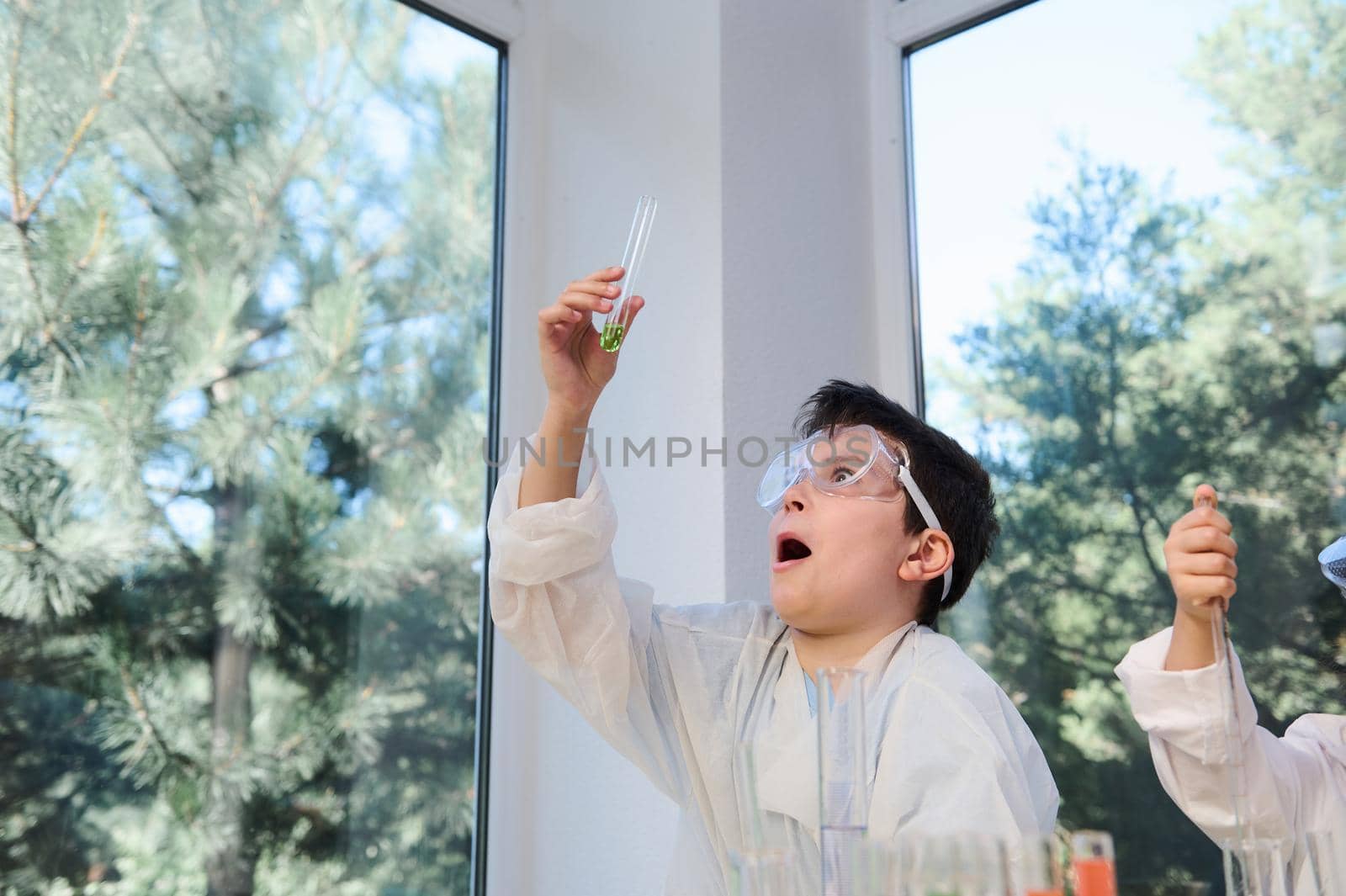 Caucasian smart nerd schoolboy in protective goggles and white lab coat holds a test tube with green chemicals and reagents and watches the taking place chemical reaction, expressing stupefaction