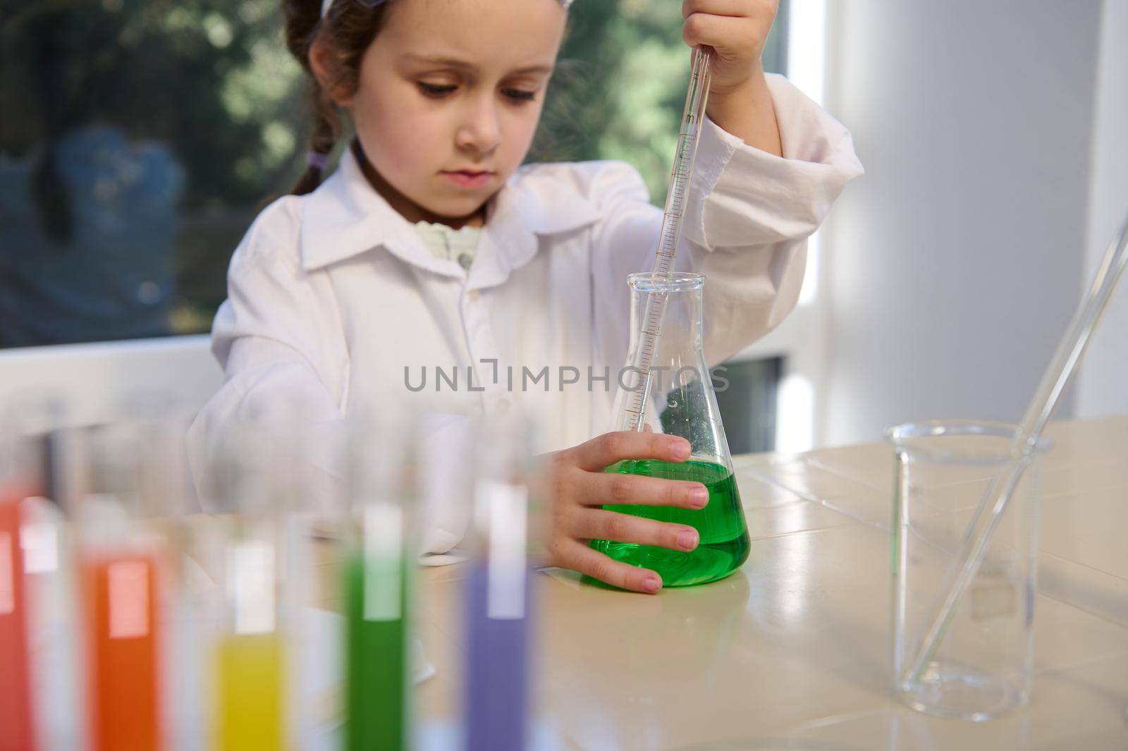 Selective focus on flat-bottomed flask with green liquid and little girl's hand holding pipette, collecting some chemicals, doing science experiments in school lab during chemistry lesson