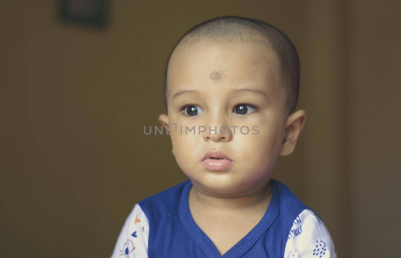 Cute bald indian baby boy in blue and white shirt looking away. Head and shoulder shot. Close up. by sudiptabhowmick