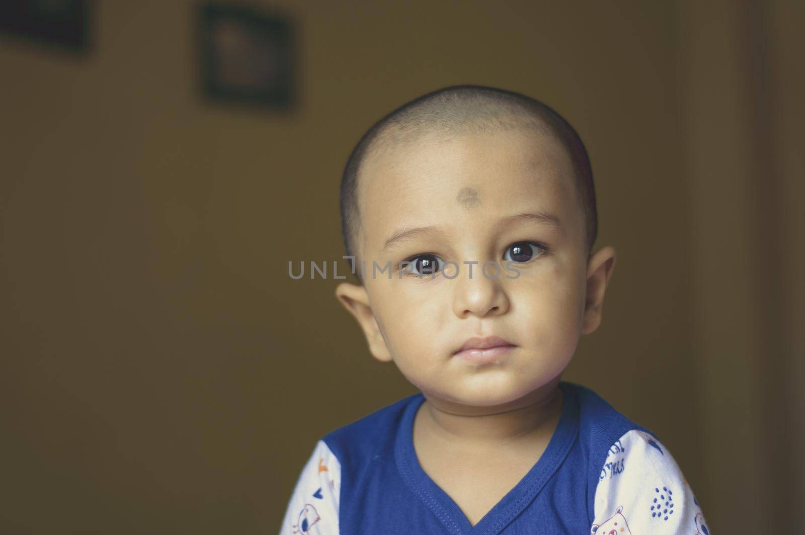 Cute bald indian baby boy in blue and white shirt looking away. Head and shoulder shot. Close up.