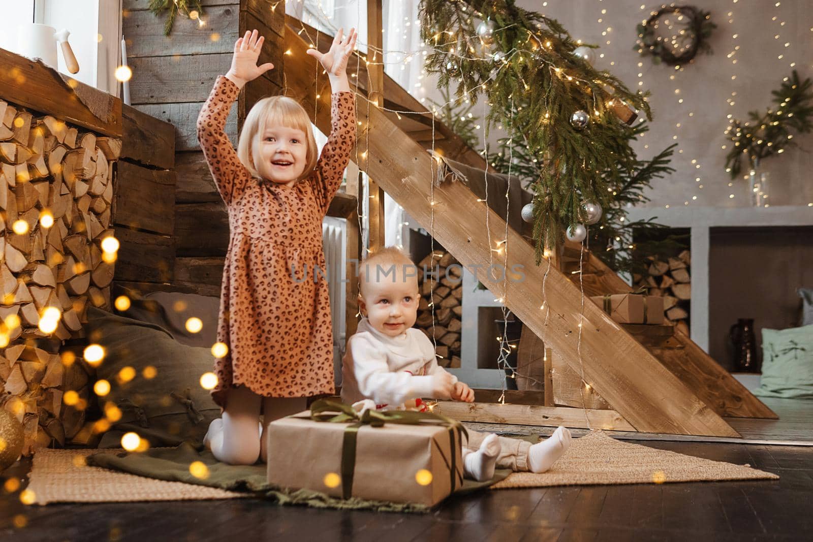Little brother and sister play on Christmas eve in a beautiful house decorated for the New Year holidays. Children are playing with a Christmas gift. Scandinavian-style interior with live fir trees and a wooden staircase.
