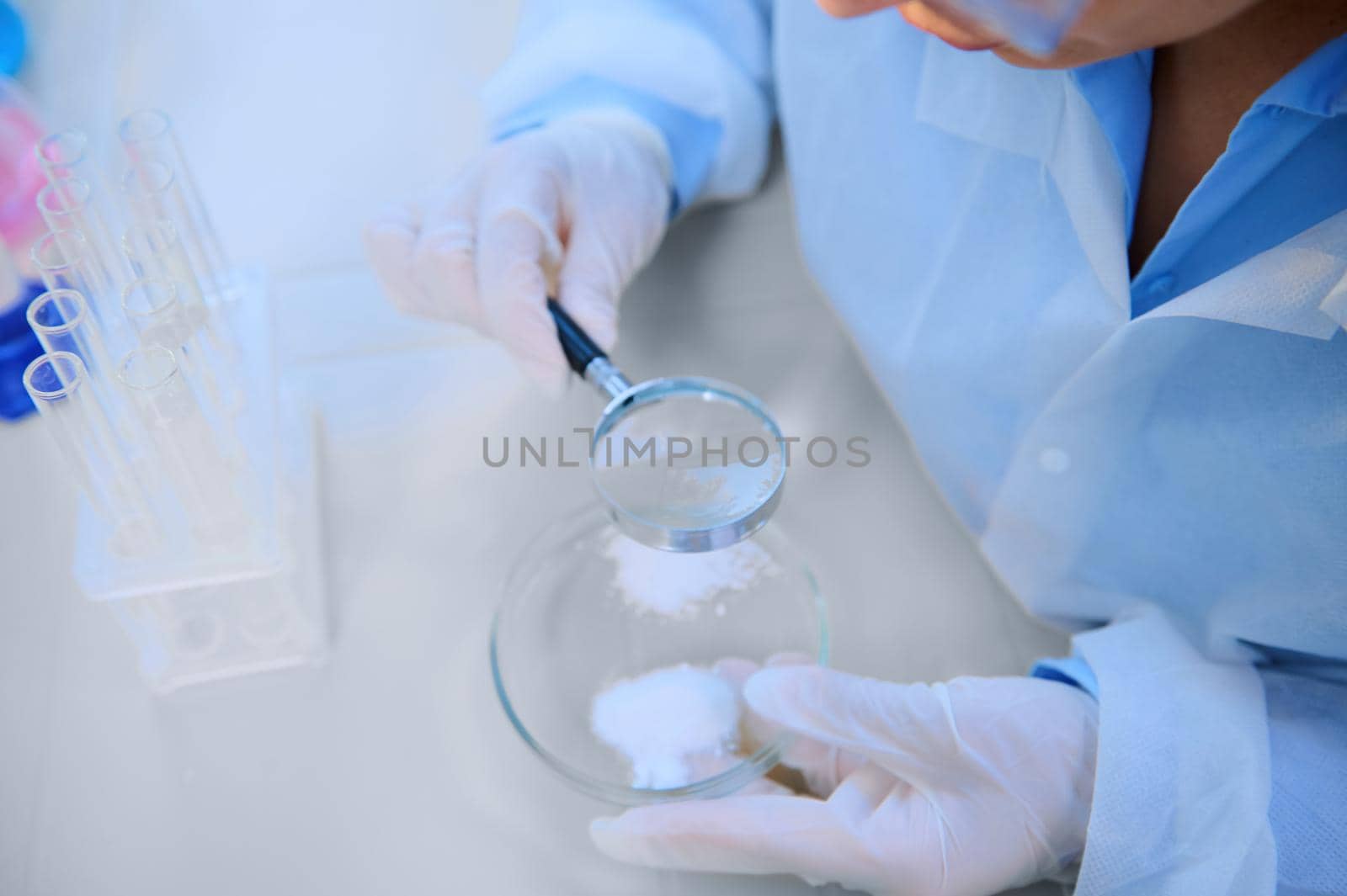 Cropped view scientist using loupe with magnifying glass to study pharmacological substance on Petri dish, sitting at table with empty test tubes on tripod in medical biolab. Investigation. Pharmacy