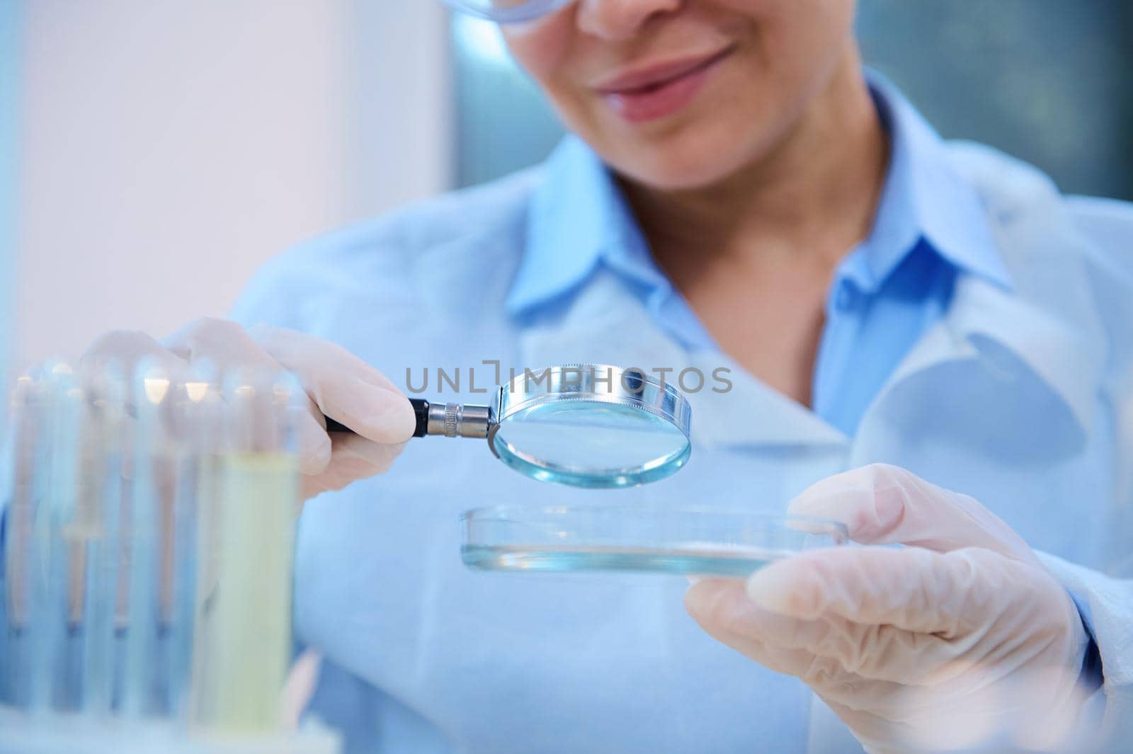 Selective focus on a loupe with magnifying glass in the hand of a woman scientist, medical biologist, examining bio material on Petri dish, while conducting experiment in science biological laboratory