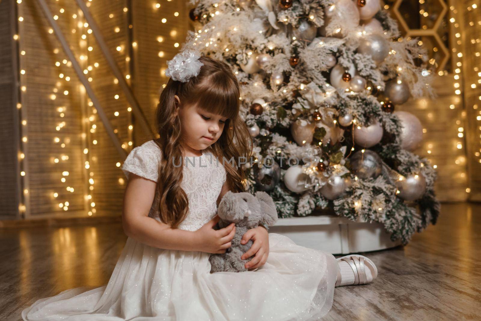 A girl in a festive light dress with a plush toy in her hands next to a Christmas tree, lights of garlands in the background. The concept of New Year holidays.