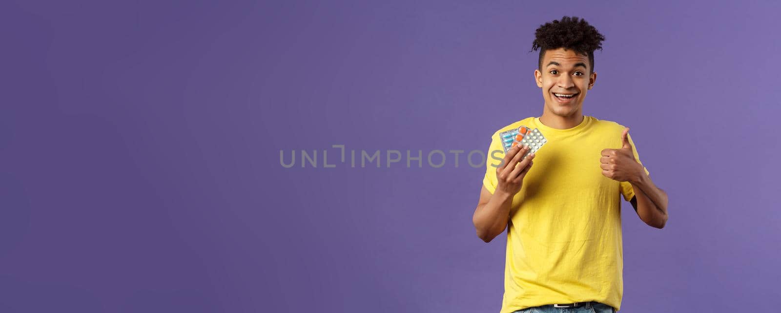 Health, influenza, covid-19 concept. Portrait of young healthy man got better, show thumbs-up holding drugs, taking pills to feel better after catching cold, being sick, purple background.