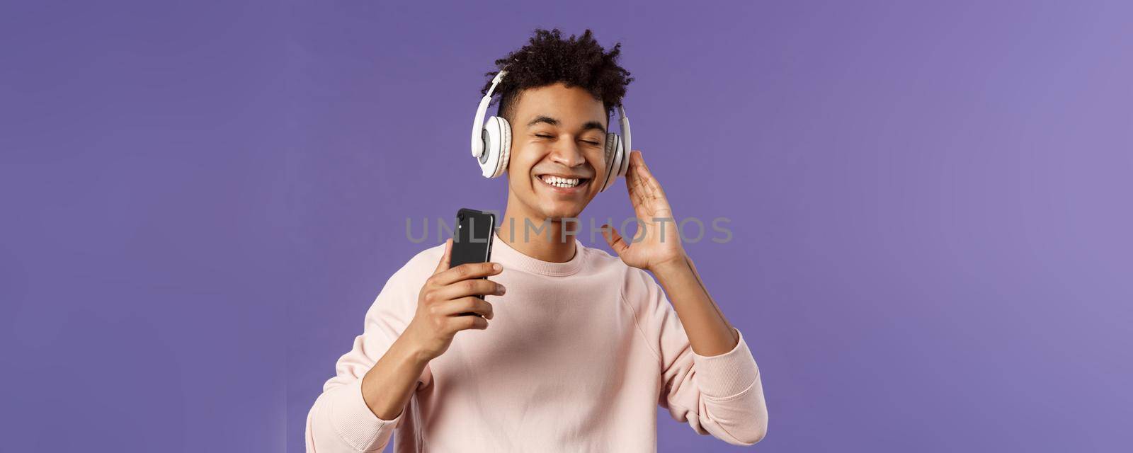 Technology and lifestyle concept. Portrait of carefree attractive young man enjoying fine sound, listening music with headphones look delighted awesome quality of bass, hold mobile phone by Benzoix