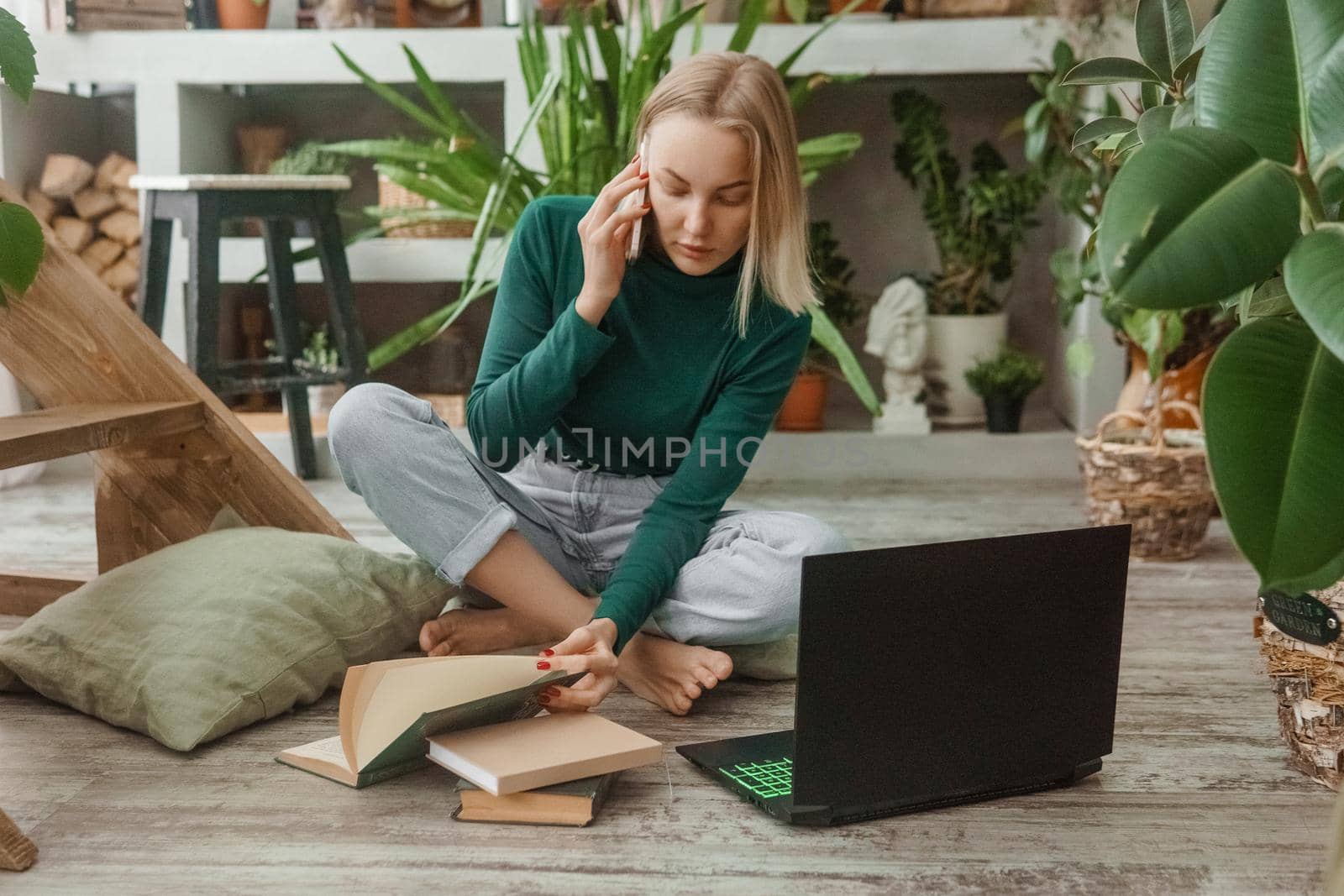 A blonde woman in a room with a lot of green indoor plants is working on a laptop. The concept of biophysical design in the interior. Work from home, work as a freelancer.