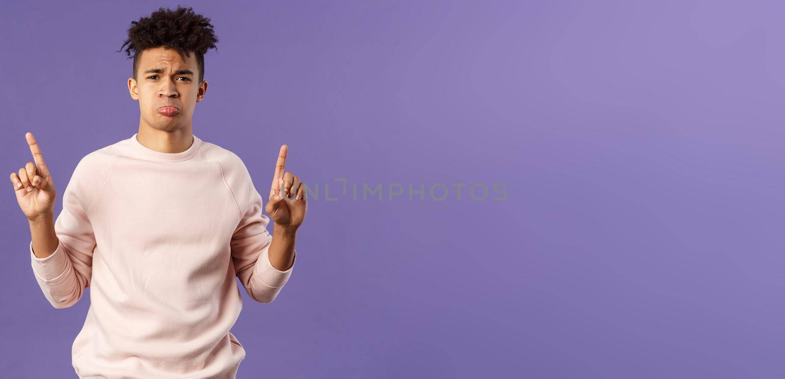 Portrait of gloomy, pouting frowning hipster guy dont have something he wants, pointing fingers up at super cool expensive thing, asking for it, trying receive compassion, purple background.
