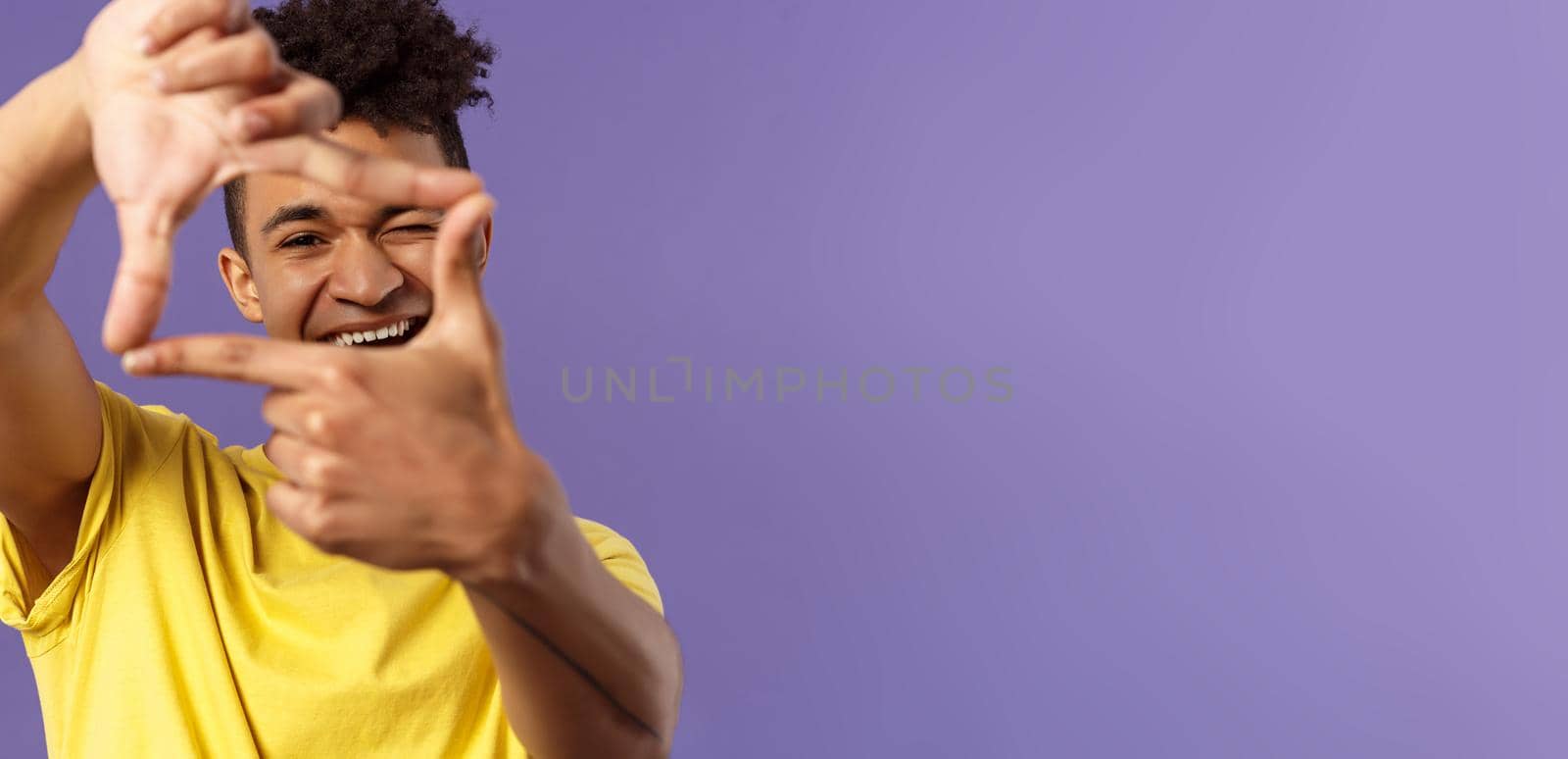 Close-up portrait of young creative photographer, man imaging, searching right angle, looking through photo frame, wink and smiling pleased, standing purple background found good shot.