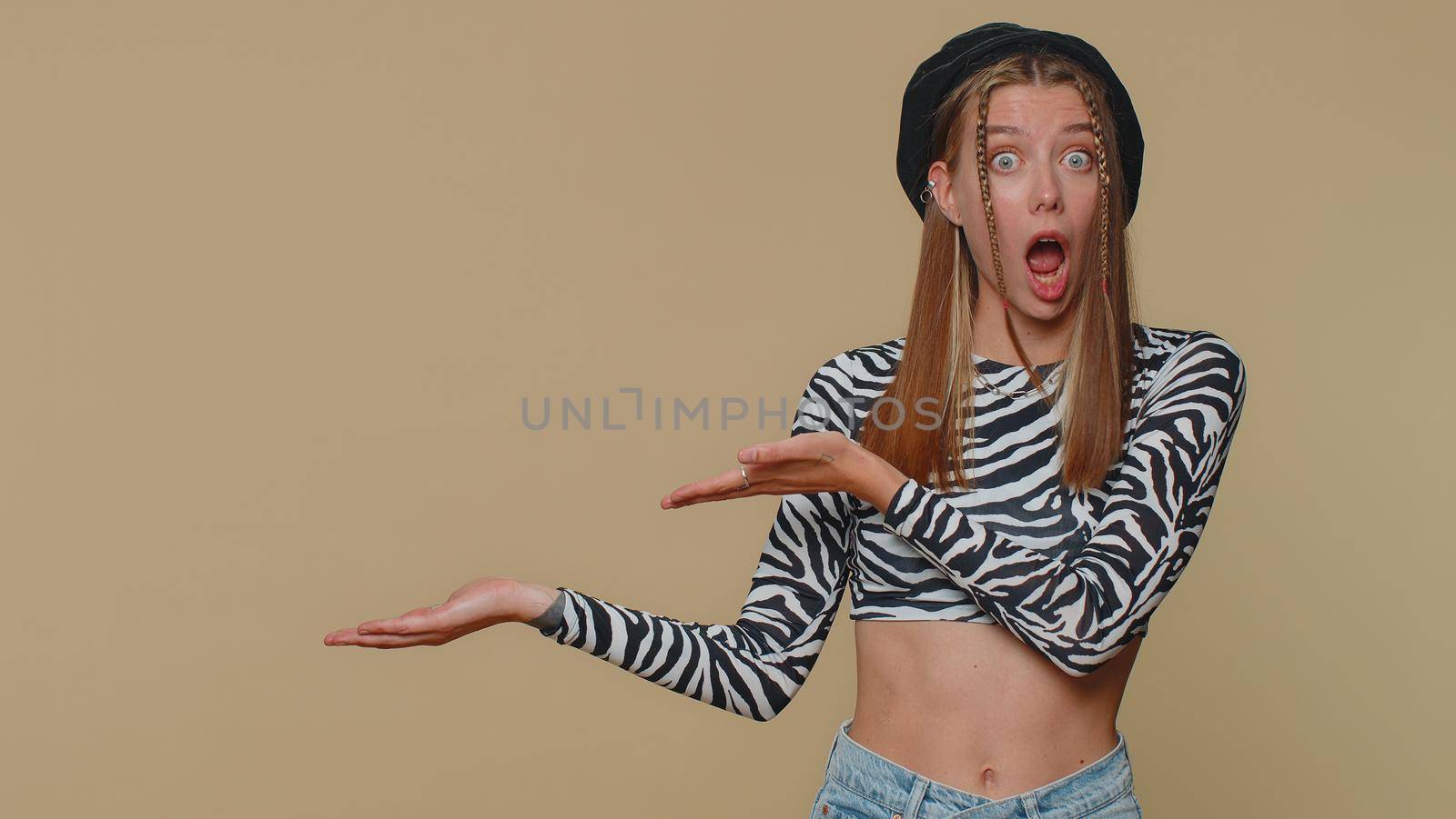 Lovely pretty young woman showing thumbs up and pointing left empty place, advertising area for commercial text, copy space for goods promotion. Adult stylish girl isolated on beige studio background